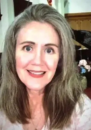 Photo of Worship Director Maura Fornataro, who has shoulder-length light brown hair and is wearing a light pink patterned shirt. She is standing indoors, with a floral arrangement and a piano in the background. The setting is Highland Presbyterian Church.